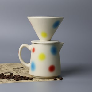 Puede incluir: Juego de vertido de café de cerámica color crema con lunares rojos, amarillos y azules. El juego incluye una jarra, una tapa y un filtro en forma de cono. Granos de café y un periódico vintage en primer plano.