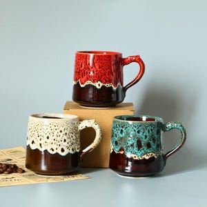 May include: Three ceramic mugs with unique designs. One is red, another is cream-colored, and the third is teal. Each mug has a brown base and a decorative handle. Coffee beans and a paper are in the foreground.