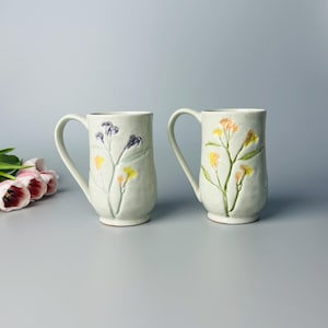 May include: Two ceramic mugs with floral designs. One mug features purple and yellow flowers, the other has yellow and peach flowers. Both mugs have a speckled, off-white finish and curved handles.