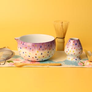 Könnte beinhalten: Ein Matcha-Teeset mit einer Keramikschale, einem Schneebesen und einer kleinen Vase, alle mit einem lila und bunten Punktmuster verziert. Enthält einen Bambuslöffel, ein Teesieb und eine Matte mit Blumenmuster. Der Hintergrund ist einfarbig gelb.