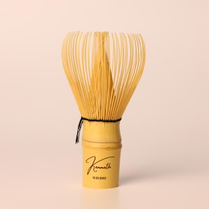May include: A bamboo matcha whisk with a light yellow handle and a bundle of thin, light yellow tines. The handle is engraved with the name "Kenneth" and the date "02.25.2025" in black script. A black string secures the tines.