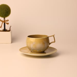 Op de afbeelding: Een goudgeel theekopje en schotelset met een uniek handvatontwerp. De kop staat op een bijpassende schotel. Een kleine, decoratieve topiary boom in een witte vierkante pot staat op de achtergrond.