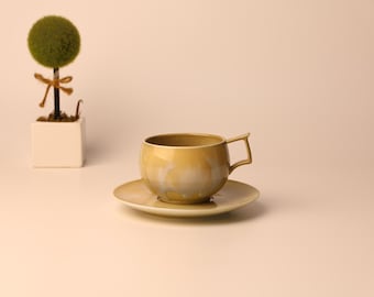 May include: A golden-yellow teacup and saucer set with a unique handle design. The cup sits on a matching saucer. A small, decorative topiary tree in a white square pot is in the background. The set is likely ceramic.