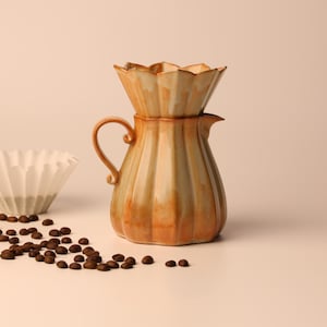 Peut inclure: Une cafetière à verser en céramique avec un dessus cannelé et une poignée, dans des tons bruns et beiges. Un filtre en papier blanc et des grains de café éparpillés sont également présents.