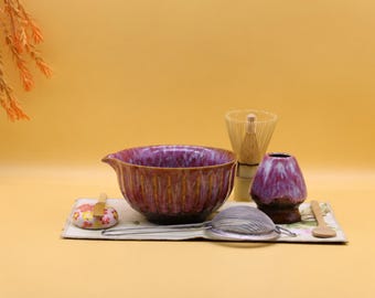 May include: A matcha tea set featuring a purple and brown glazed ceramic bowl with a pouring spout, a bamboo whisk, a purple ceramic whisk holder, a metal mesh strainer, a small wooden spoon, and a floral patterned ceramic rest.