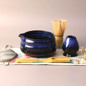 May include: A matcha tea set with a blue ceramic bowl, whisk, and a small vase. The set includes a bamboo whisk, a wooden scoop, and a metal sieve, all arranged on a floral patterned cloth.