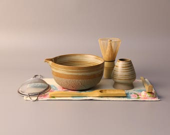 May include: A matcha tea set with a ceramic bowl, whisk, and tea caddy. The bowl is brown and tan with a textured exterior. A bamboo scoop and whisk are also included, along with a metal sieve and a floral patterned cloth.