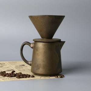 Può includere: Set per versare il caffè color bronzo con un filtro a forma di cono sopra una brocca abbinata. Il set è su un giornale d'epoca con chicchi di caffè sparsi. La brocca ha un manico e un beccuccio.