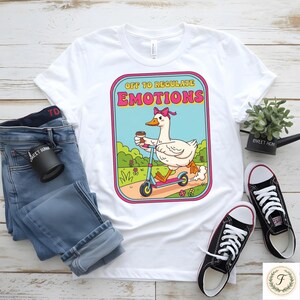 May include: White t-shirt with a graphic of a goose riding a scooter, holding a coffee cup, and wearing a pink bow. The text on the shirt reads "OFF TO REGULATE EMOTIONS". The shirt is paired with blue jeans and black sneakers.