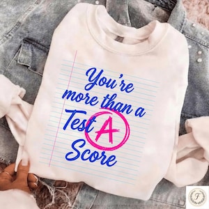 May include: White sweatshirt with the text "You're more than a Test A Score" in blue script. The "A" in "Test A Score" is inside a pink circle. The sweatshirt is on top of a denim jacket.