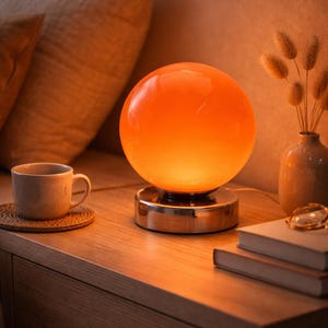 Op de afbeelding: Een oranje bolvormige tafellamp met een chromen voet staat op een houten nachtkastje. Een kopje koffie op een onderzetter, een vaas met droogbloemen en boeken zijn ook aanwezig. De lamp geeft een warm licht.
