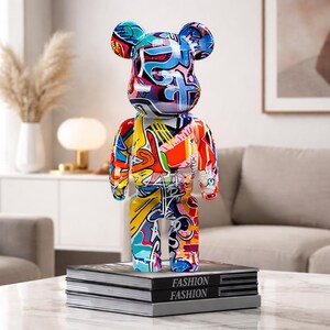 May include: A colorful, abstract art bear figurine with a graffiti-style design. The bear is standing on a stack of books with the word "FASHION" printed on them. The bear's design includes blue, red, yellow, and white colors.