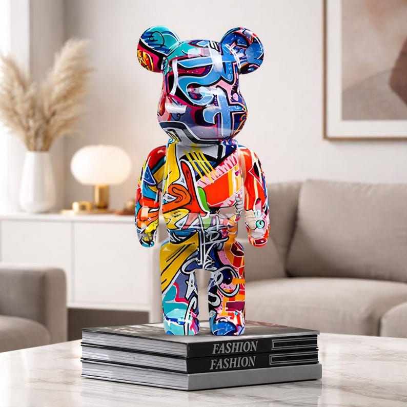 May include: A colorful, abstract art bear figurine with a graffiti-style design. The bear is standing on a stack of books with the word "FASHION" printed on them. The bear's design includes blue, red, yellow, and white colors.