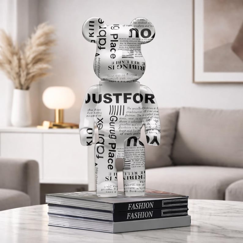 May include: A white and gray decorative bear figurine with text, standing on a stack of books labeled "FASHION." The bear has a glossy finish and features newspaper-style text. The figurine is a modern home decor item.