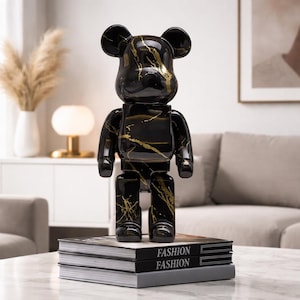 May include: A black and gold decorative bear figurine with a marble-like pattern. The figurine is standing on a stack of books with the word "FASHION" printed on the spines. The bear has a glossy finish and a modern design.