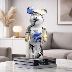May include: A white bear-shaped figurine with abstract brushstroke designs in blue, black, yellow, and gray. The figurine is standing on a stack of books with the word "FASHION" printed on the spine.