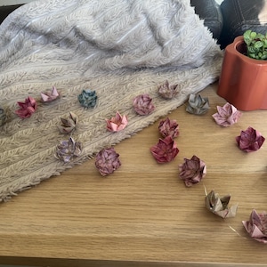 Puede incluir: Una guirnalda de luces con flores de loto de origami en varios tonos de rosa, morado, gris y azul. Las flores de papel están conectadas por un cable fino con una pequeña batería blanca en un extremo, dispuestas sobre una superficie de madera.