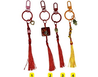 May include: Four keychains with decorative charms and tassels. The keychains have red, pink, and orange metal hardware. The charms include a heart, a square with an Om symbol, an owl, and a Hamsa hand. The tassels are red and yellow.