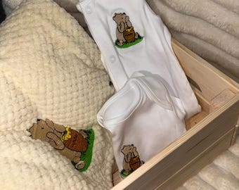 Könnte beinhalten: Ein weißer Babybody und ein weißer Baby-Strampler, beide mit einem gestickten beigen Bären, der einen Honigtopf mit der Aufschrift "HUNNY" hält, in einer hellen Holzkiste neben einer strukturierten, cremefarbenen Decke platziert.
