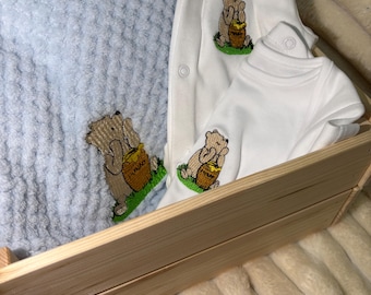 Könnte beinhalten: Eine hellblaue, strukturierte Decke und zwei weiße Baby-Bodys mit einer Stickerei eines Bären, der einen Honigtopf mit der Aufschrift HUNNY hält. Die Artikel sind in einer hellen Holzkiste auf einem weichen, cremefarbenen Stoff angeordnet.