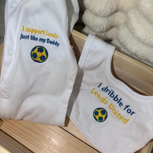 Puede incluir: Conjunto de bebé blanco con body y babero. El body tiene el texto "I support Leeds just like my Daddy" y un balón de fútbol. El babero dice "I dribble for Leeds United" con un balón.