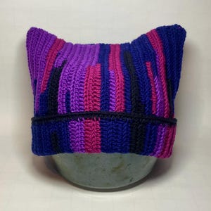 Könnte beinhalten: Eine handgefertigte Häkelmütze mit Katzenohren. Die Mütze hat ein gestreiftes Muster in Lila-, Rosa- und Blautönen mit schwarzen Akzenten. Der Rand ist einfarbig dunkelblau. Die Mütze wird auf einer hellgrünen Schale präsentiert.