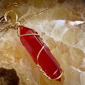 May include: A faceted, deep red, hexagonal crystal pendant wrapped in thin gold-colored wire. The crystal is suspended from a delicate gold-colored metal chain. The pendant features a pointed tip and a smooth, polished surface with a vibrant red hue.