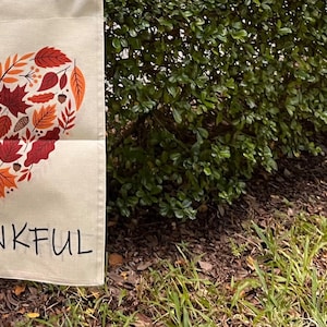 May include: A beige garden flag with a heart-shaped design made of fall leaves in shades of red, orange, and brown. The word "THANKFUL" is printed in black block letters below the heart. The flag is displayed outdoors.