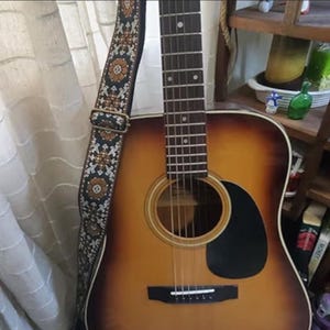 May include: Acoustic guitar with a sunburst finish, featuring a black pickguard and a decorative guitar strap with a floral pattern in brown, tan, and white against a black background. The guitar is leaning against a white curtain.