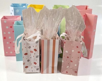 May include: A collection of colorful gift bags in various patterns, including polka dots, terrazzo, and stripes. Some bags are filled with tissue paper and tied with white ribbon. The bags are in shades of pink, blue, yellow, and green.
