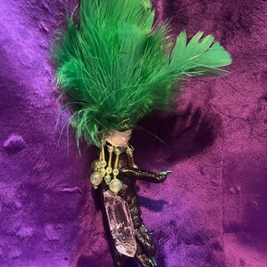 May include: A handcrafted, whimsical accessory featuring a black, claw-like base adorned with a clear crystal and dangling beads. The design is topped with vibrant green feathers, creating a unique and artistic piece.