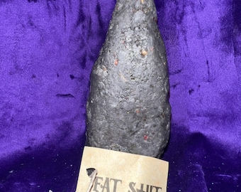 Op de afbeelding: Een donkergrijze, getextureerde kaars met een witte lont, tegen een paarse achtergrond. Een klein, bruin label aan de kaars leest "EAT SHIT" in zwarte hoofdletters. De kaars heeft een ruwe, organische vorm.