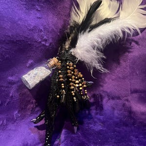 May include: A handcrafted, black, claw-like decorative item with gold-colored beads and white and black feathers. A small glass bottle with a cork stopper is attached. The item is set against a purple, textured background.
