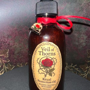 May include: A brown glass bottle with a black dropper and a red ribbon tied around the neck. The bottle features a label reading "Veil of Thorns Ritual Protection Oil." A small gold charm with a red rose is attached to the ribbon.