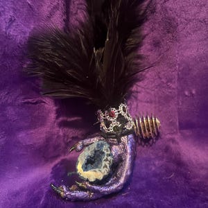 May include: A fantasy-themed sculpture featuring a purple dragon claw holding a geode. The claw is adorned with a crown and black feathers. A gold-colored spiral element is attached to the claw. The sculpture is set against a purple background.