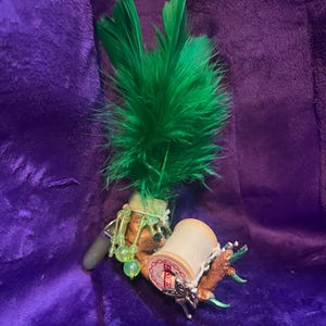 May include: A handmade sewing-themed accessory with a green feather accent. It features a wooden spool of thread, a small charm, and decorative beads. The accessory is set against a purple background.