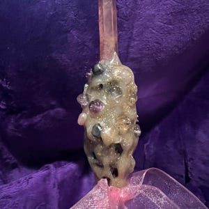 Op de afbeelding: Een handgemaakte toverstaf met een grote, roze kristallen punt. De handgreep van de toverstaf is versierd met diverse edelstenen en glitter, en is afgewerkt met een roze lint. De toverstaf staat tegen een paarse achtergrond.