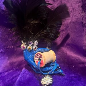 Op de afbeelding: Een grillig, handgemaakt ambachtelijk stuk met zwarte veren, een blauwe stoffen basis en een kleine kroon. Het bevat een houten garenklos met de tekst "Talon Mercury Sewing Fast" en een hanger met een draadkooi-ontwerp. Paarse achtergrond.