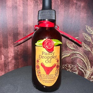 Könnte beinhalten: Eine braune Glasflasche mit einer schwarzen Pipette und einem roten Band um den Hals gebunden. Auf dem Etikett steht "Panty Dropper Oil" mit einer Grafik roter Unterwäsche. Das Etikett enthält auch den Text "For Seduction, Confidence & Carnal Mischief, Wicked Waters Witchery."