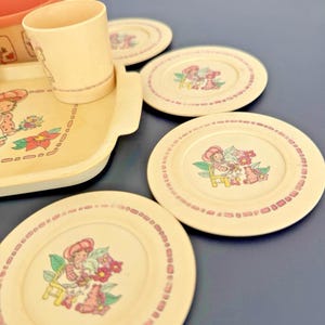 May include: A vintage children's tea set in cream and pink. The set includes a tray, cup, plates, and a container. Each piece features a cartoon character with flowers and a pink dotted border.
