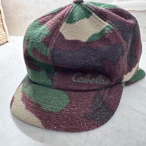 Peut inclure: Une casquette de baseball en polaire avec un motif camouflage dans des tons vert foncé, bordeaux et beige. Le panneau avant présente le mot Cabela's brodé en fil vert foncé. La casquette possède une calotte arrondie et une visière incurvée.