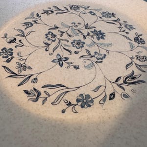 Op de afbeelding: Een ronde, gebroken witte plaat met een blauw bloemmotief. Het patroon toont gestileerde bloemen en bladeren in een cirkelvormige opstelling. De plaat heeft een gespikkelde textuur en een blauwe rand.