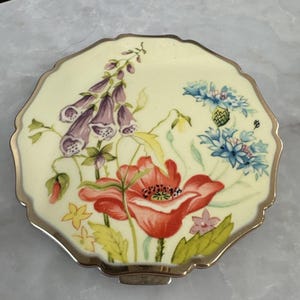 May include: A round, scalloped-edge compact mirror with a cream-colored enamel top featuring a painted floral design. The illustration includes purple foxgloves, a large red poppy, and small blue cornflowers with green leaves and stems.