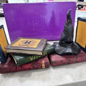 May include: A collection of decorative items featuring a wizarding theme. Includes a black Sorting Hat, several faux books with gold-colored pages, and a purple box in the background. The books are inscribed with words like "Hogwarts" and "Potions".