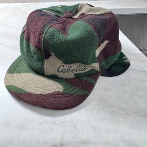 Peut inclure: Une casquette de baseball en polaire avec un motif camouflage dans des tons de vert, marron et beige. Le panneau avant affiche le mot Cabela's brodé en fil vert foncé. La casquette présente une calotte arrondie et une visière incurvée.