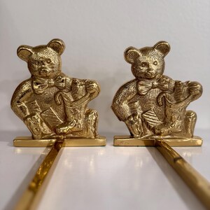 May include: Two gold-colored teddy bear figurines. Each bear is seated, wearing a bow tie, and holding a bag. The bears are attached to a rectangular base with a long, thin rod extending from the base. The background is a plain white surface.