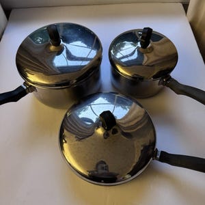 Può includere: Un set di tre pentole in acciaio inossidabile con coperchi e manici neri. Le pentole sono di diverse dimensioni, con una superficie lucida e riflettente. I coperchi hanno pomelli neri. Le pentole sono disposte su una superficie bianca.
