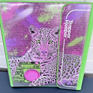 May include: A neon green and purple Trapper Keeper binder featuring a leopard with a boombox. The leopard is pink and white with green eyes. The background has a brick wall design with purple and green paint splatters. The Mead logo is in the upper left corner.