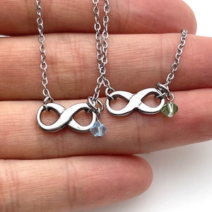 May include: Two silver infinity symbol necklaces with small crystal charms. The necklace on the left has a light blue charm, and the necklace on the right has a green charm.