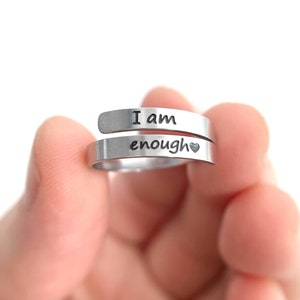 May include: A silver ring with the words "I am enough" engraved on it. The ring is held in a hand with a red sweater sleeve visible.